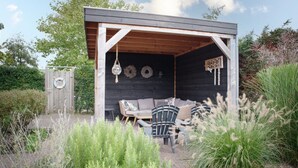 A covered outdoor seating area with a wooden bench and chairs, surrounded by greenery.