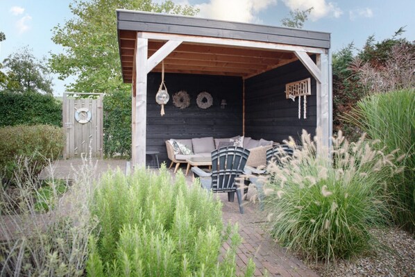 A covered outdoor seating area with a wooden bench and chairs, surrounded by greenery.