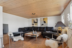 A modern living room with a wooden ceiling, black leather sofa, a glass coffee table, and a white fluffy chair. - Cozy holiday home in Harboøre (Harboøre)