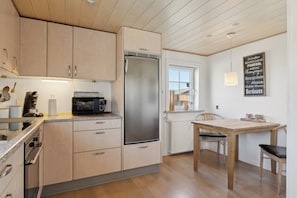 A modern kitchen with wooden cabinets, a stainless steel refrigerator, a microwave, and a dining table with chairs. - Cozy holiday home in Harboøre (Harboøre)