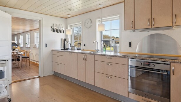 A modern kitchen with wooden cabinets, a stainless steel oven, and a dining area with a table and chairs. - Cozy holiday home in Harboøre (Harboøre)