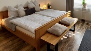 Una cama doble con estructura de madera, ropa de cama gris y varias almohadas. Una mesita de noche con una lámpara y un cuadro colgado en la pared.