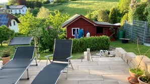 Un patio con dos sillones de descanso, un pequeño edificio rojo y un perro al fondo.