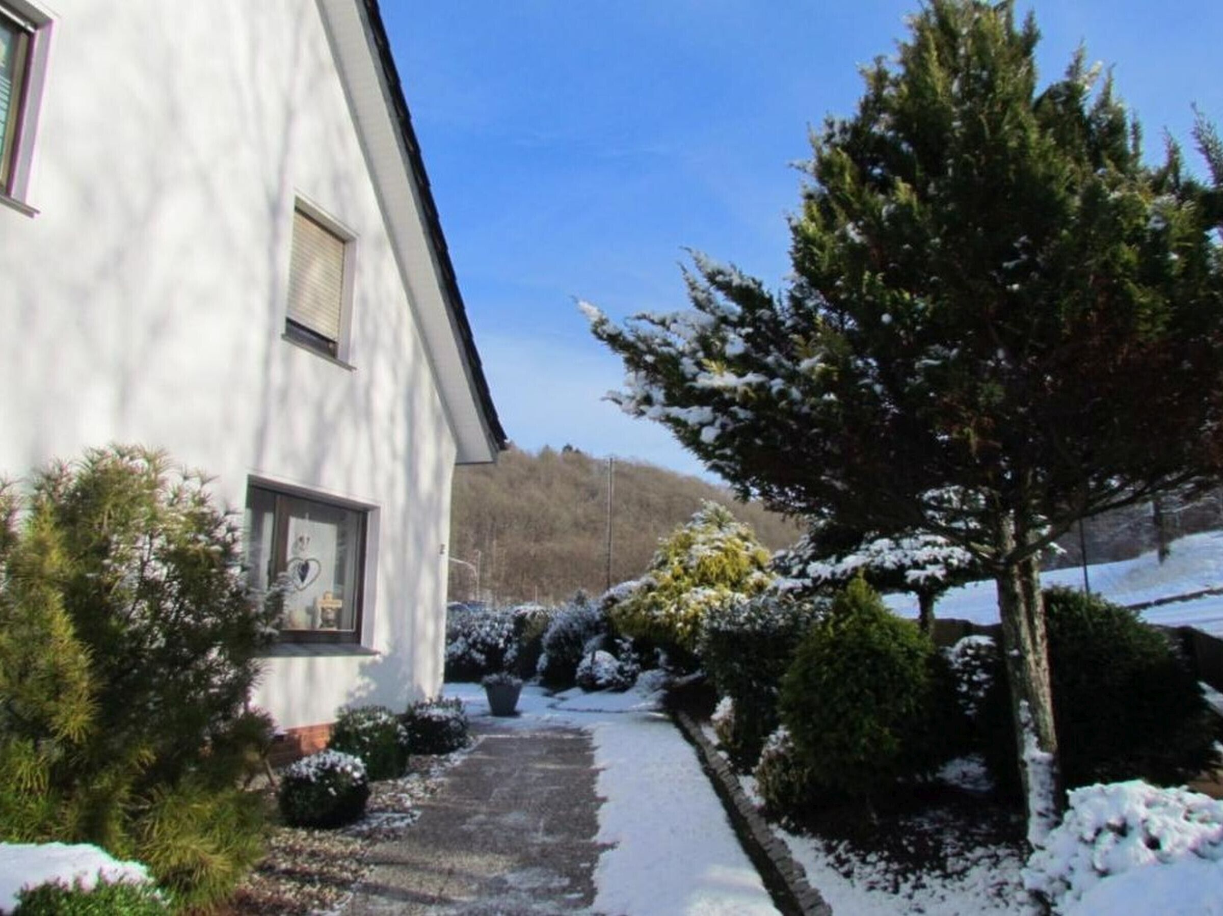 A white building with a visible window and door, surrounded by snow-covered trees and shrubs.