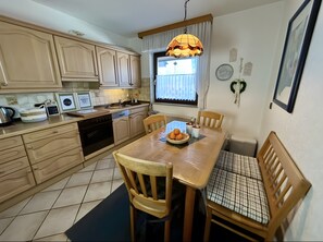 A kitchen with wooden cabinets, a dining table with chairs, a hanging light fixture, and a window with curtains. - Holiday Home with Garden, Terrace and Wi-Fi (Sundern)