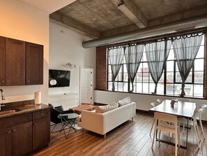 Un espacio habitable compacto con kitchenette, un sofá, una mesa de comedor y un amplio ventanal con cortinas.