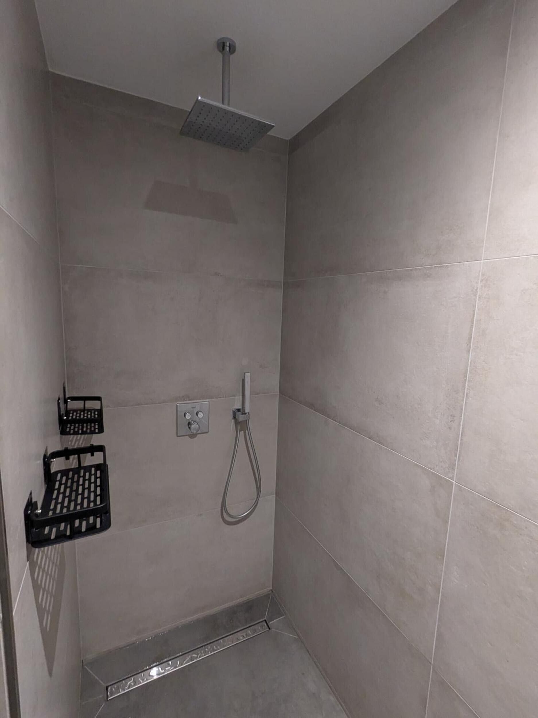 Een douche met een handdouche, een bedieningspaneel aan de muur, en een zwart plankje met toiletartikelen.