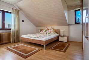 Une chambre avec un plafond en pente, un lit avec une literie à motifs floraux, une table de chevet, une commode et un radiateur.