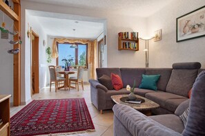 A living room with a sofa, a coffee table, a dining table with chairs, a red patterned rug, a bookshelf, and a window with curtains. - Apartment 'Plocher' with Lake View, Shared Garden and Wi-Fi (Immenstaad am Bodensee)