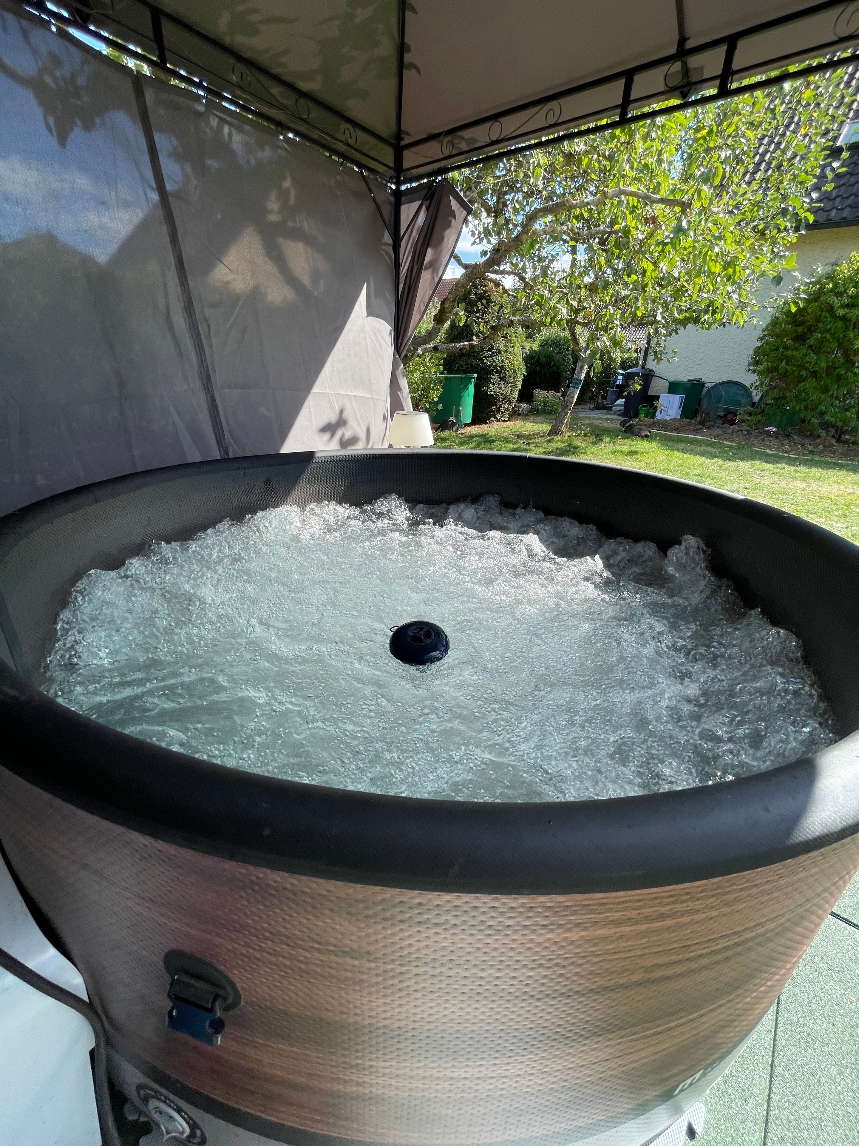 A large outdoor hot tub with a black rim and a textured surface, surrounded by a covered area with a metal frame.