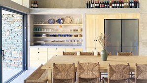A modern kitchen with a dining area, featuring a large wooden table, wicker chairs, and a wine rack above the counter. - Villa Mariana by Boutique (Folegandros)