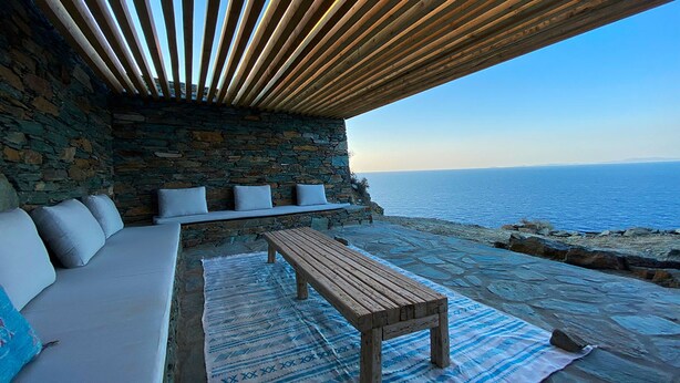An outdoor seating area with a stone wall, wooden bench, and cushions. - Villa Mariana by Boutique (Folegandros)