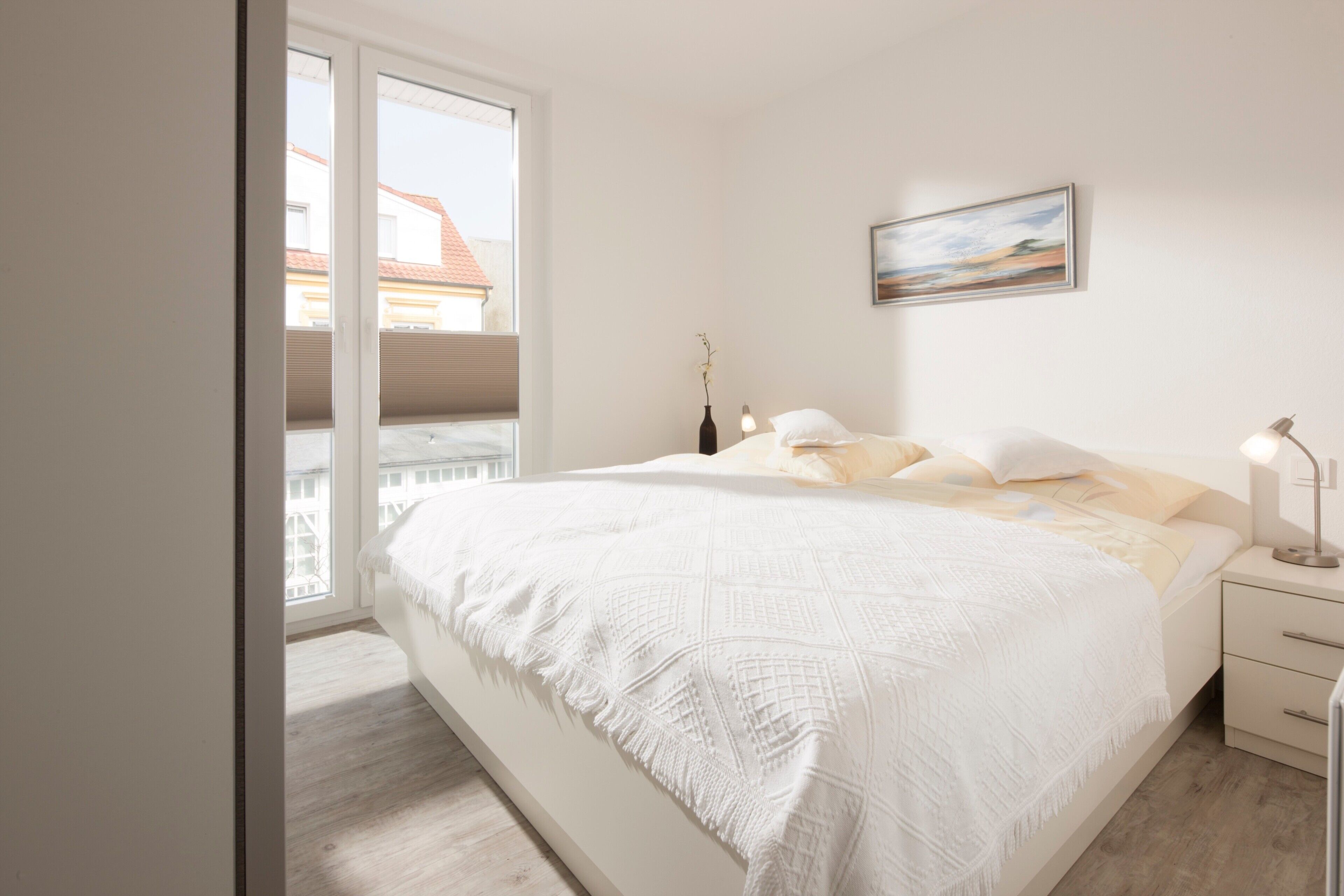Ein Schlafzimmer mit einem großen Bett, einem Nachttisch, einer Lampe, einem Gemälde und Blick auf Gebäude durch eine Schiebetür.