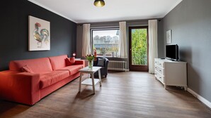 A modern living room with a red sofa, a white coffee table, a grey armchair, a TV on a white cabinet, and a large window with curtains. - Apartment 'Ostseeblick Fewo 6' with Private Terrace, Balcony and Wi-Fi (Kronsgaard)