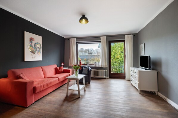 A modern living room with a red sofa, a white coffee table, a grey armchair, a TV on a white cabinet, and a large window with curtains.