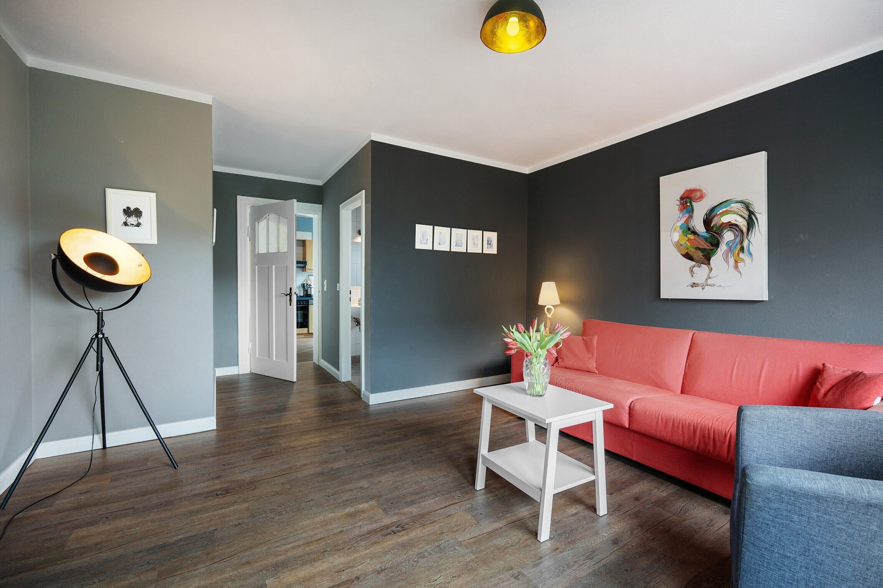 A modern living room with a red sofa, a white coffee table, a tripod, and a framed rooster painting on the wall.