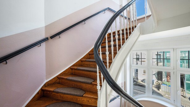 A staircase with wooden steps and a black handrail, leading to a landing with a view of the outside.