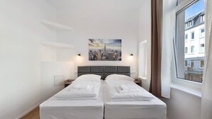 A double bed with a cityscape painting above it, a window with curtains, and a wooden floor.