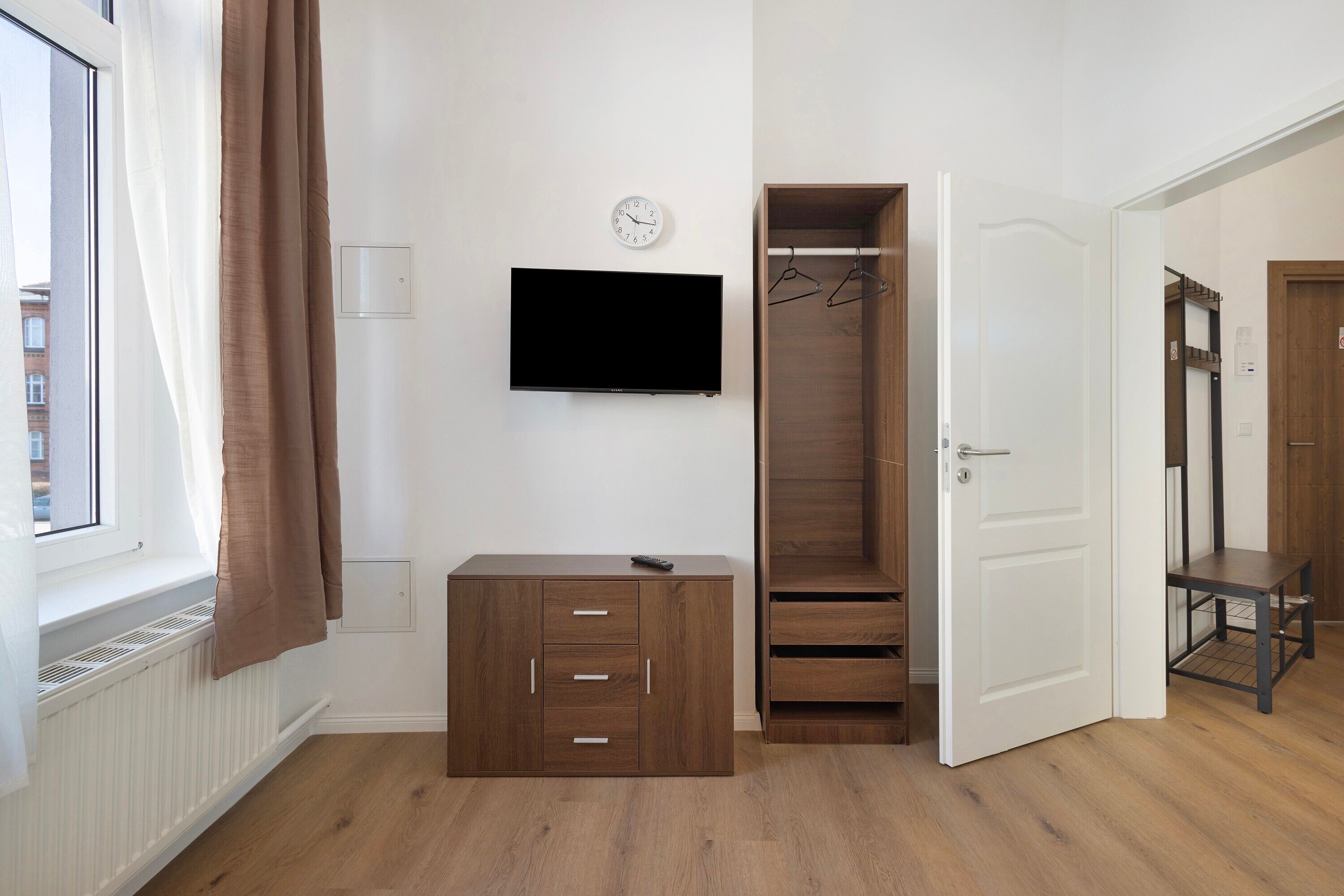 A room with a window, a TV mounted on the wall, a wooden cabinet, a wardrobe, and a door leading to another room.