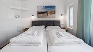 A double bed with white linens, a headboard, a wall-mounted clock, a beach scene painting, and a window with blinds.