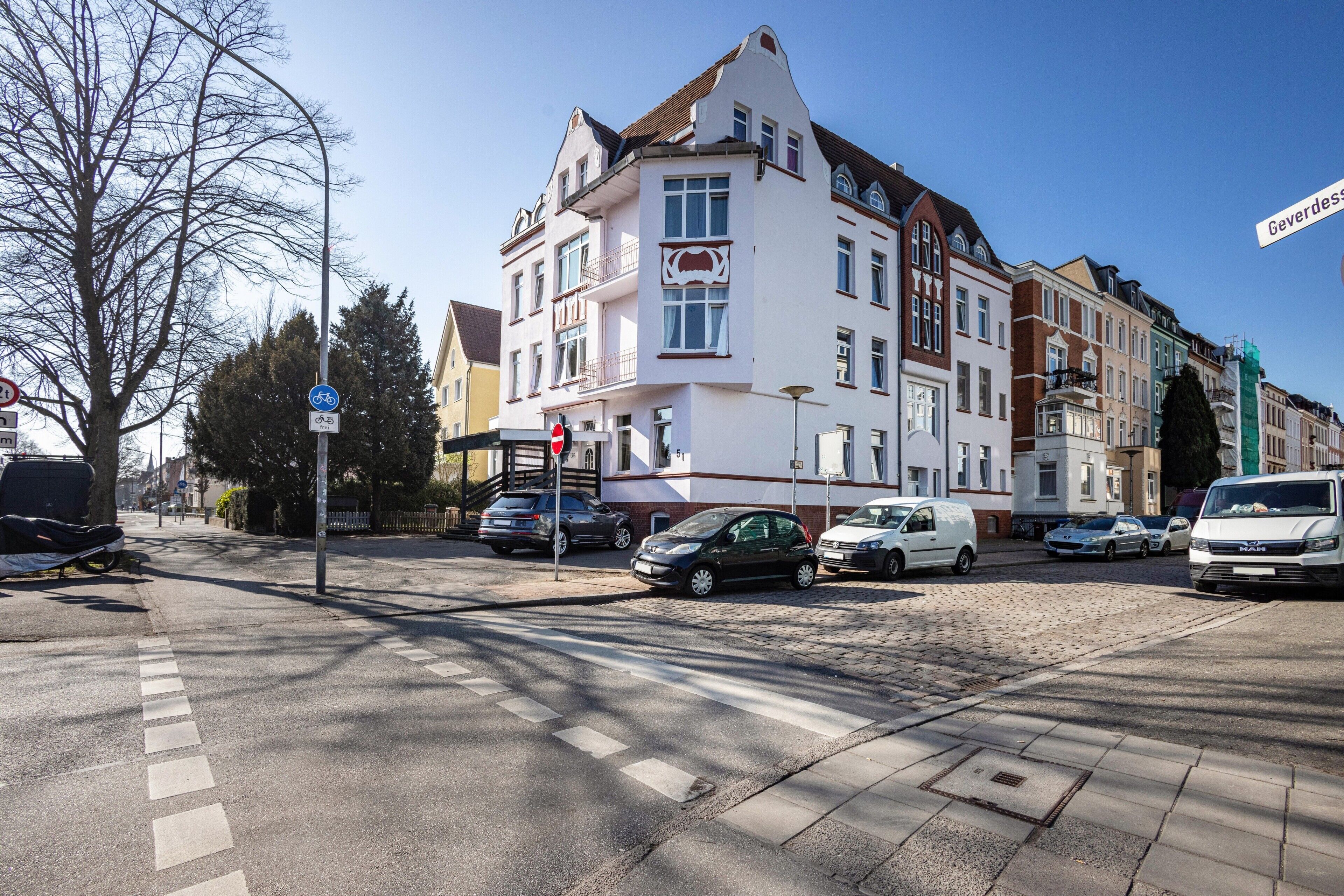 Een straat met geparkeerde auto's, een zebrapad en een rij meerverdiepingen met een mix van bouwstijlen.