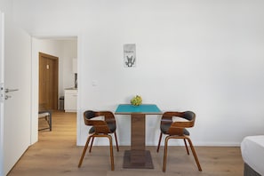 Una habitación con suelo de madera, una mesa con dos sillas, una mesita con frutero y una puerta que da a otra habitación.