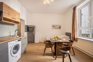 A compact kitchen with a washing machine, a dining area with a table and chairs, and a large window with curtains.