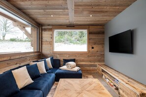 A modern interior with a wooden ceiling, a large window, a dark blue sofa, a flat-screen TV, and a wooden coffee table.