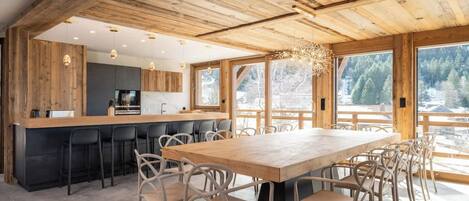 Eine moderne Küche mit Holzboden, ein Essbereich mit großem Tisch und Stühlen sowie große Fenster mit Blick auf Bäume.