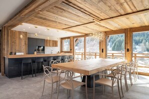 Une cuisine moderne avec un plafond en bois, un coin repas doté d’une grande table et de chaises, et de grandes fenêtres offrant une vue sur les arbres.