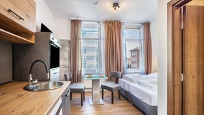A compact hotel room with a kitchenette, a dining area, a bed, and a view of the city.