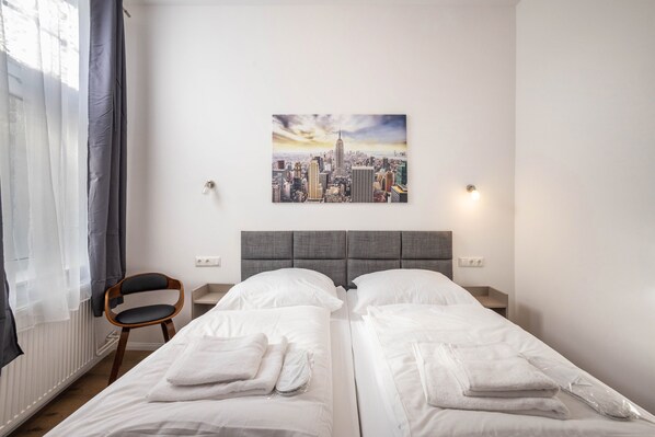 A hotel room with two beds, a cityscape painting on the wall, a chair, and a window with curtains.