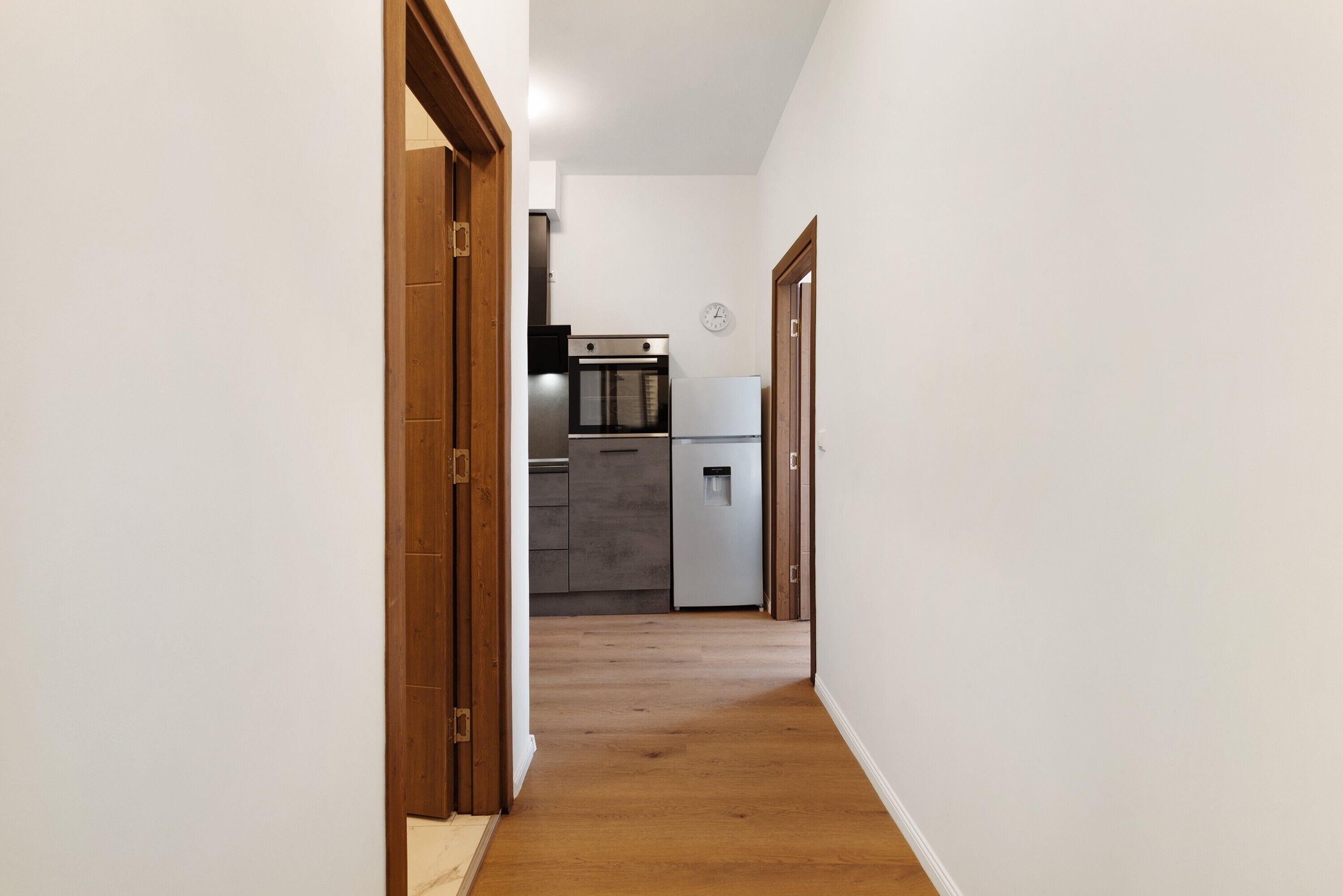 A narrow hallway with wooden doors and a kitchen area in the background.