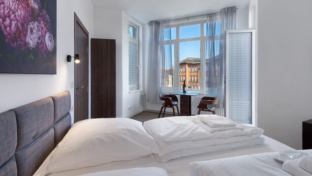 A hotel room with a bed, a desk, and a view of buildings through large windows. - 2-bedroom apartment 3-2 with balcony and Wi-Fi (Lübeck)