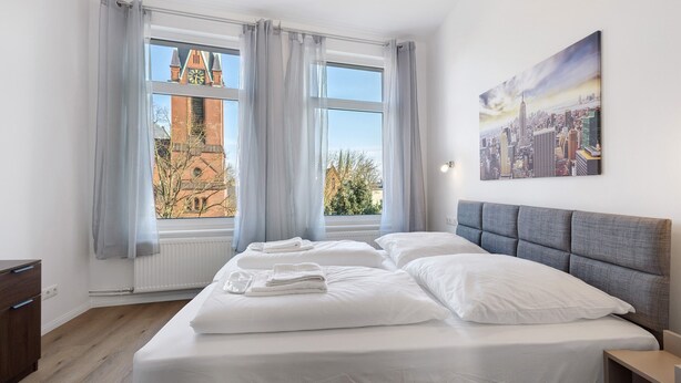 Een hotelkamer met een groot bed, een hoofdbord, een raam met gordijnen, een radiator, een houten dressoir en een stadsgezichtschilderij aan de muur.
