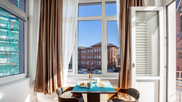 A room with a table, chairs, a window with curtains, and a view of buildings. - 2-room apartment 2-2 with balcony and Wi-Fi (Lübeck)