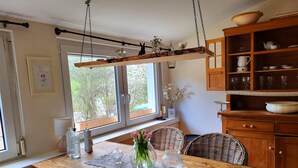 Een houten eettafel met een kantonderzetter, rieten stoelen, een hanglamp en een raam met uitzicht op bomen.