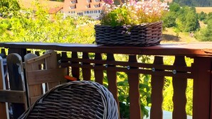 Een houten terras met een rieten stoel, een rieten mandje en uitzicht op een groen landschap met gebouwen.