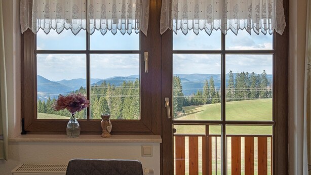 Ein Zimmer mit großem Fenster, einer Blumenvase und einer hölzernen Bank.