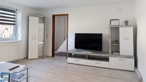 A modern living room with a flat-screen TV, built-in shelving, a glass coffee table, and a view through a window with blinds. - Holiday Home 'Ferienhaus' with Private Terrace, Private Garden and Wi-Fi (Zernitz-Lohm)