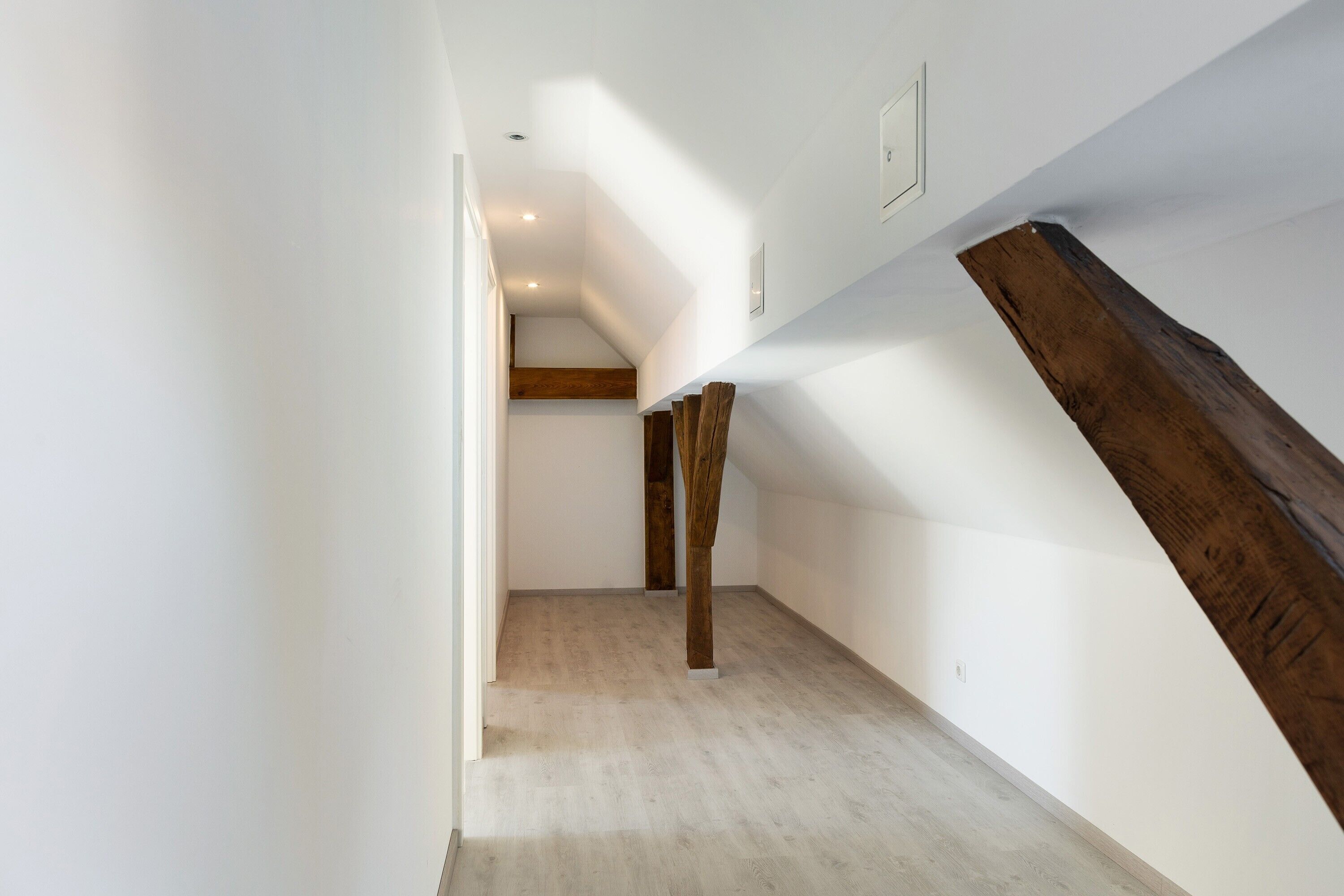 Un couloir étroit avec une poutre en bois, des murs blancs et un plancher de couleur claire.