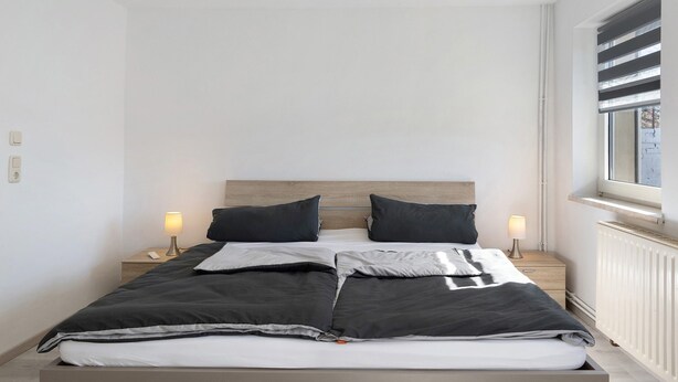 A modern bedroom with a wooden headboard, a bed with black and white bedding, two bedside lamps, and a window with blinds. - Holiday Home 'Ferienhaus' with Private Terrace, Private Garden and Wi-Fi (Zernitz-Lohm)