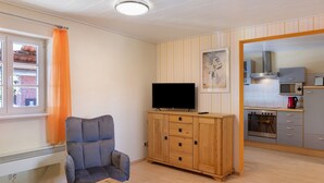 A living room with a television, a wooden cabinet, a chair, and a kitchen area with appliances. - Studio '30 sqm' with Wi-Fi in Historic Four-Sided Farmhouse (Friedewald)