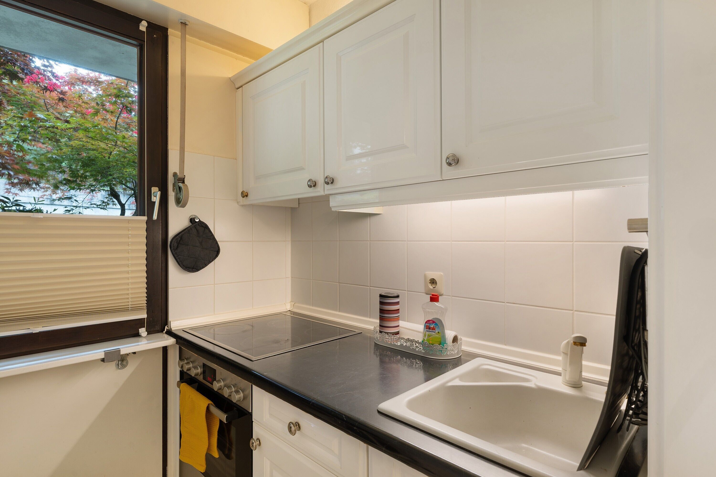 A kitchen with white cabinets, a black countertop, a window with blinds, and a view of trees outside.