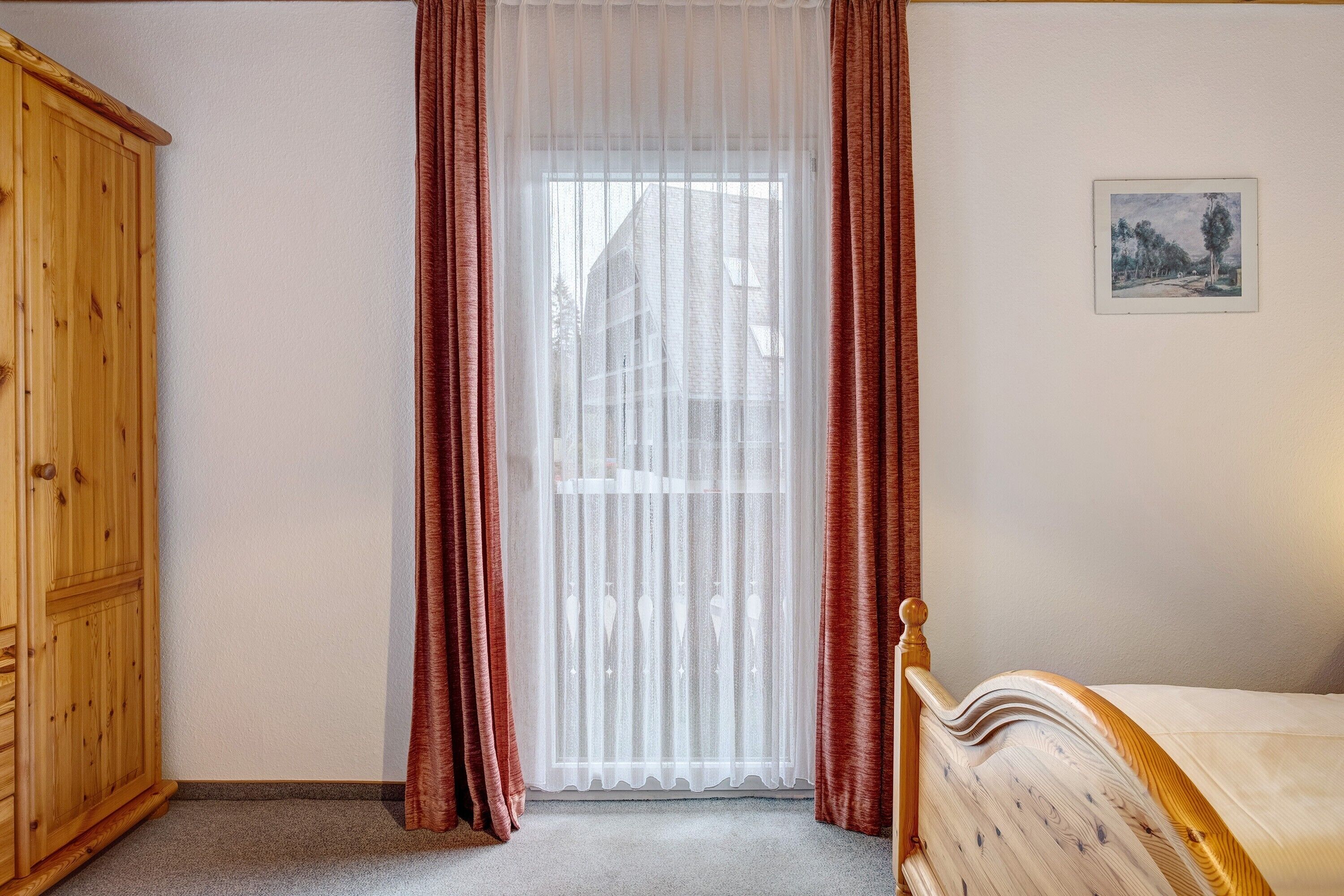Une chambre avec une armoire en bois, un lit avec une tête de lit en bois, une fenêtre avec des rideaux légers et un tableau encadré accroché au mur.