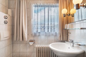 Een badkamer met een witte wasbak, een zeepdispenser, een toiletpapierhouder, een raam met een kantelaar en een radiator.