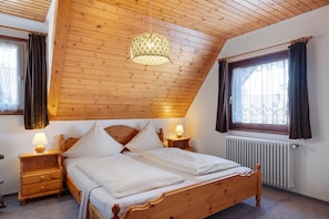 Une chambre avec un plafond en bois, un lit recouvert de draps blancs, une table de chevet avec une lampe et une fenêtre avec des rideaux.
