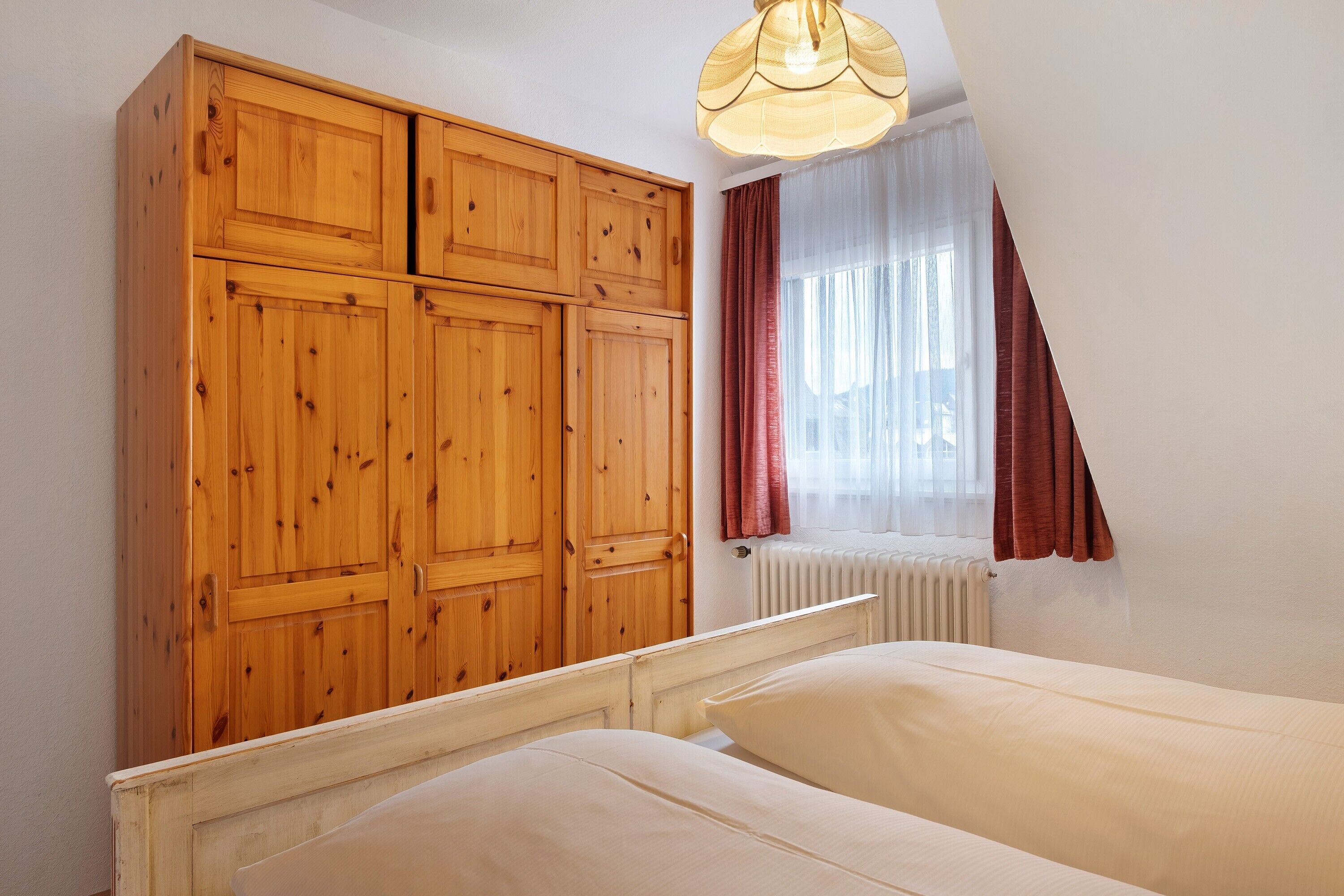 A bedroom with a wooden wardrobe, a bed, and a window with curtains.