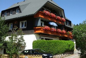 Een gebouw met twee verdiepingen, inclusief een balkon op de tweede etage, een auto ervoor en een haag op de achtergrond.