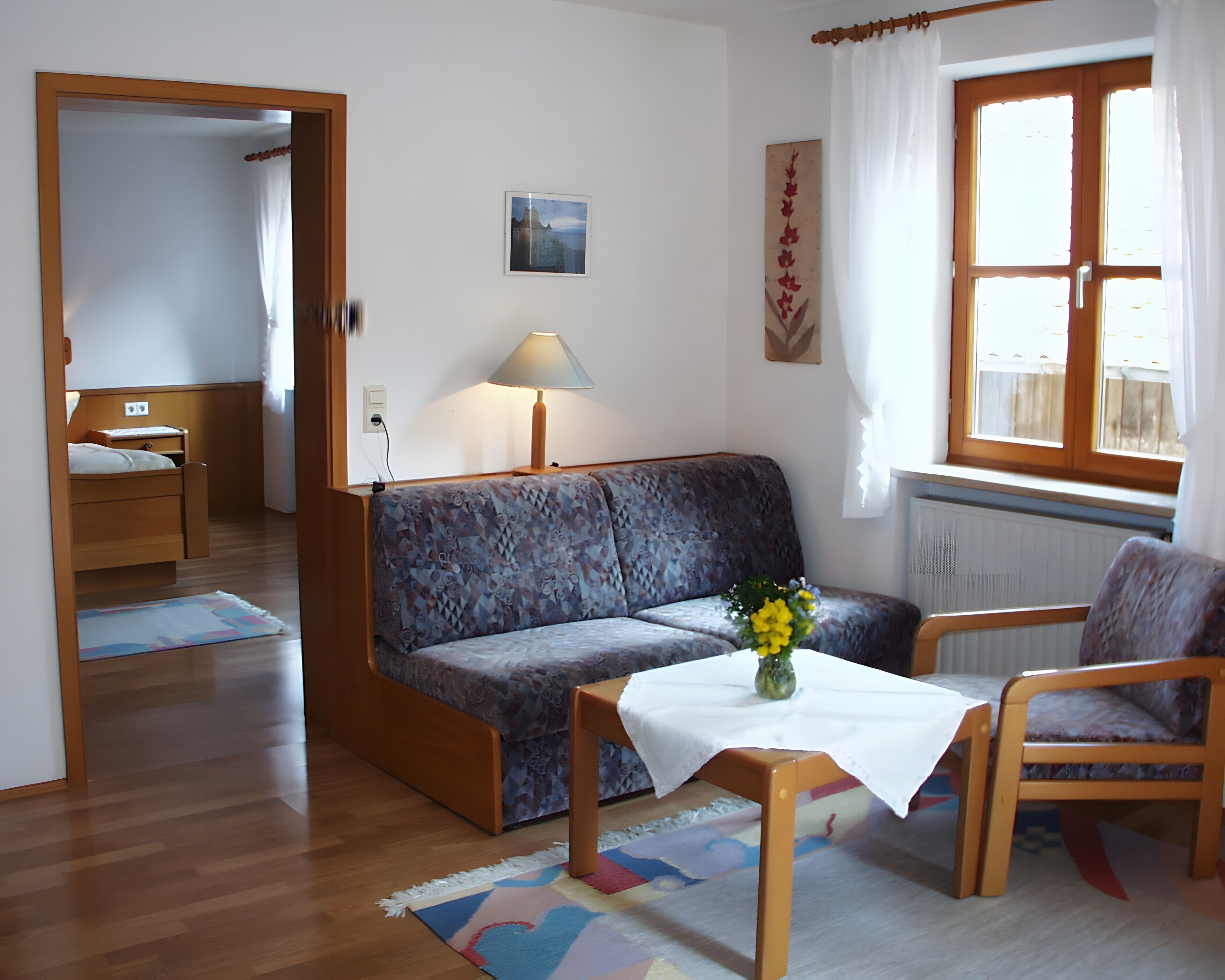 Ein Zimmer mit einer Couch, einem Tisch mit einer Vase, einem Stuhl, einem Bett, einem Fenster mit Vorhängen und einem Heizkörper.